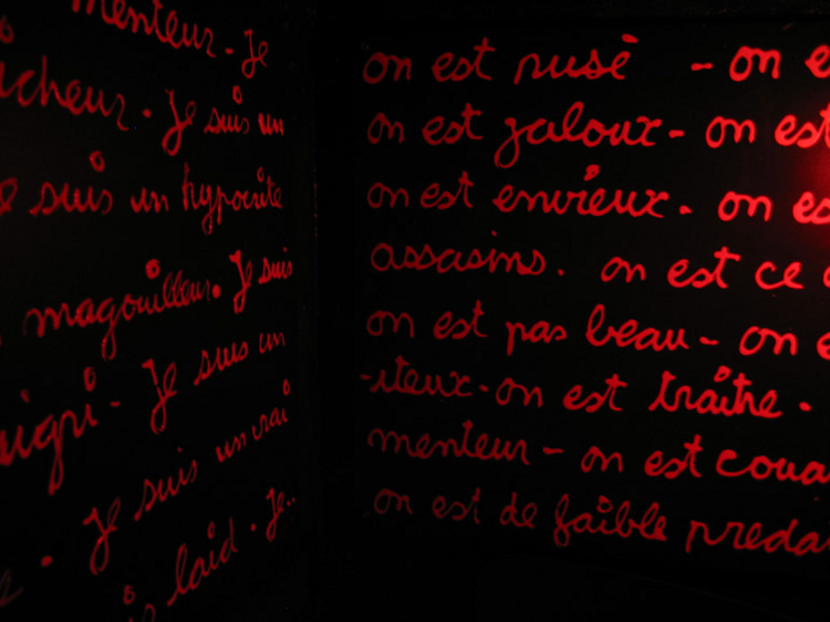 L'interno di una *cabina* di legno nera. Le tre pareti sono piene di scritte rosse sul nero e sono illuminate da una lampadina rossa. Noi vediamo una parte di parete sinistra e una parte di quella centrale, in prospettiva. La luce viene da destra.